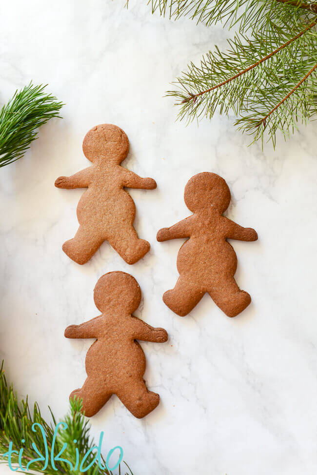Three gingerbread men on a white marble background surrounded by fresh evergreen branches