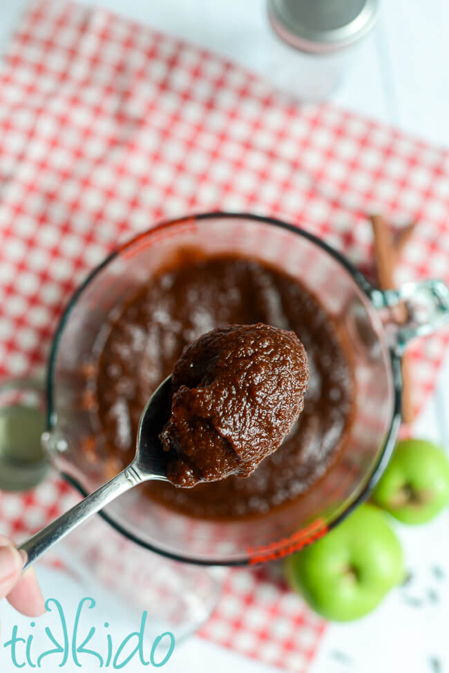 Spoon holding thick homemade apple butter above a pyrex measuring cup holding more apple butter.  The pyrex cup measure is sitting on red and white gingham cloth, and surrounded by fresh green apples and cinnamon sticks.