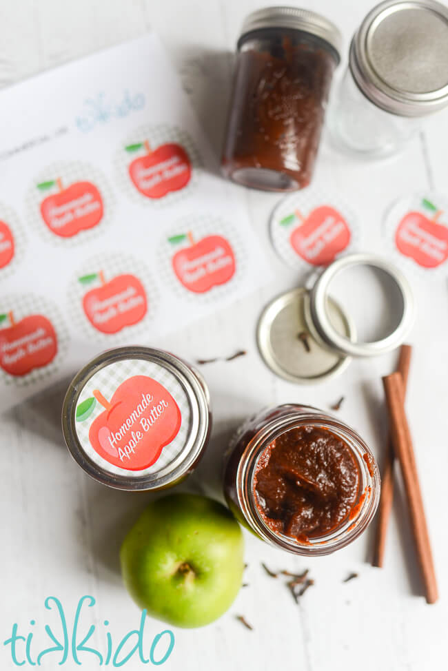 Jar of Instant Pot Apple Butter next to a sheet of printable canning lid labels reading "homemade apple butter."  Apples, whole spices, and extra canning lids are scattered around.