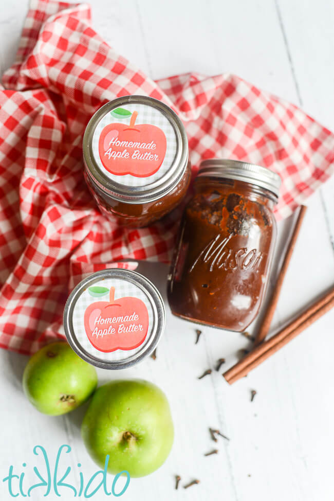 Instant Pot Apple Butter Recipe in three jars, surrounded by fresh apples, cinnamon sticks and cloves, and a red and white checkered kitchen towel.