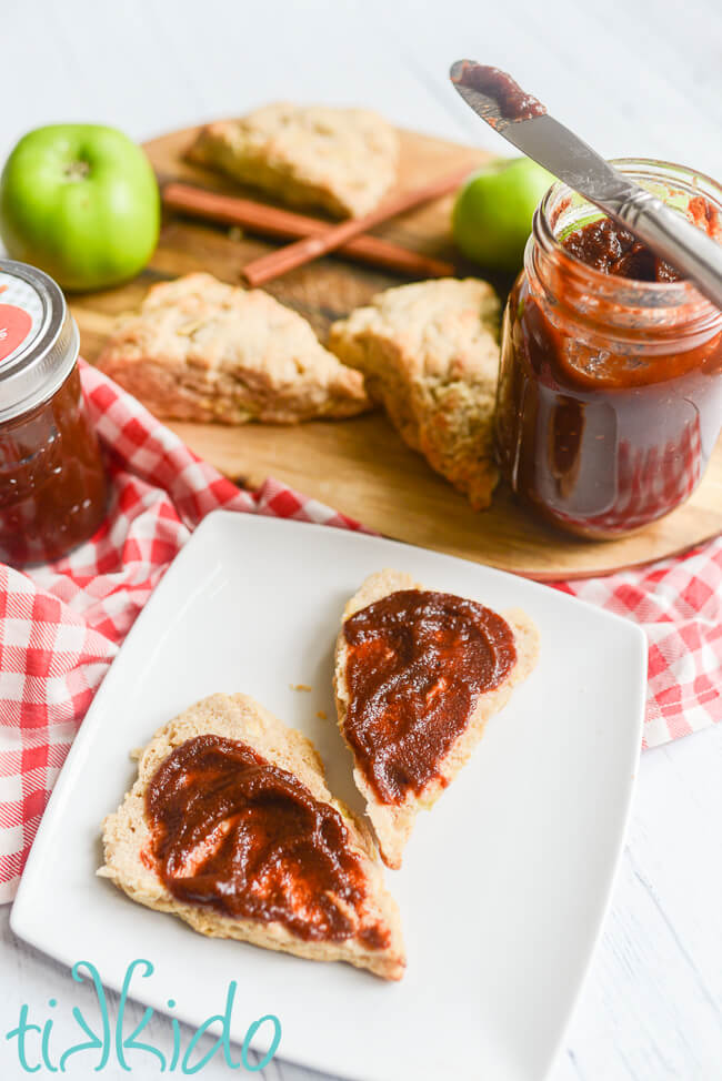 Apple butter spread on apple scones, in front of a cutting board with a jar of apple butter, apple scones, fresh apples, and cinnamon sticks.