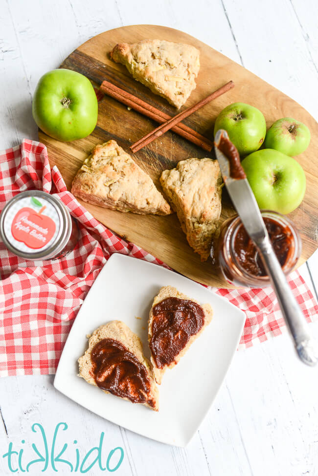 Cutting board with apple scones, apples, and apple butter, next to a plate with an apple scone spread with homemade apple butter.