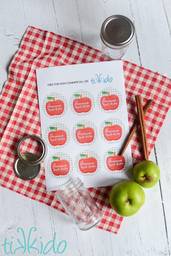 Printable canning lid labels reading "homemade apple butter" with a green and white gingham pattern with a red apple graphic.  The paper is on a red and white chekered cloth, and surrounded by empty canning jars, apples, and whole spices.