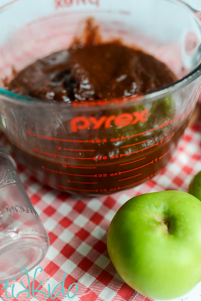 Photo showing reduced Instant Pot Apple Butter in a pyrex cup measure, surrounded by green apples and empty canning jars.