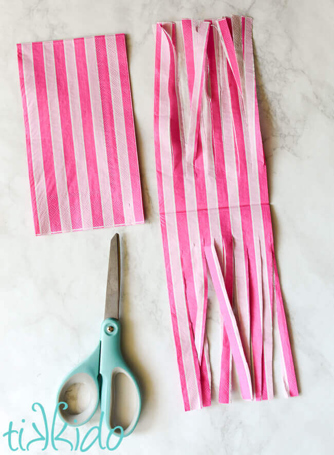 Two pink striped paper napkins, one folded next to a pair of scissors, one unfolded and cut into thin ribbons.