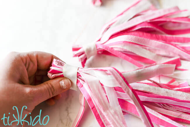 White ribbon being wrapped around the top of a striped pink napkin tassel.