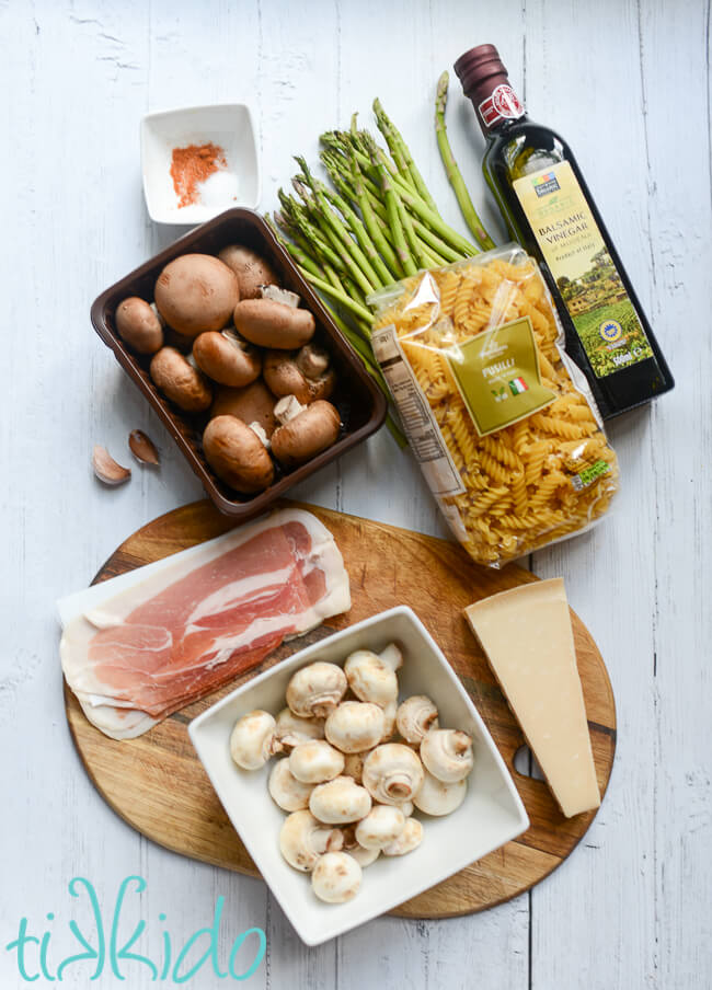 Ingredients for prosciutto, asparagus, and mushroom pasta recipe on a white wooden surface.