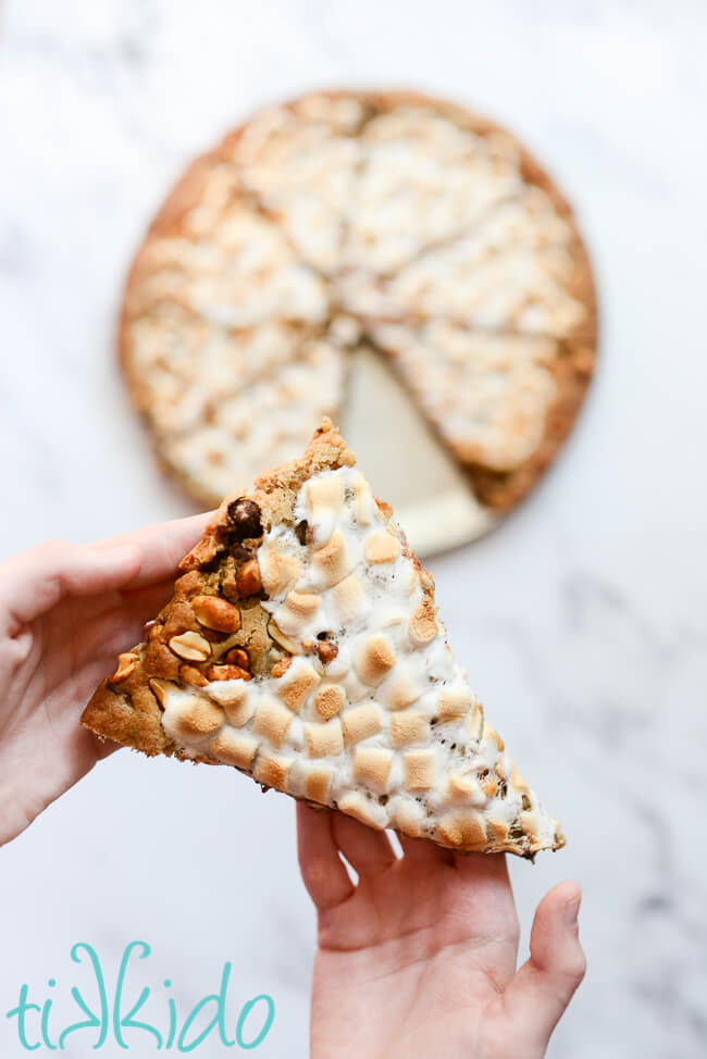 Two hands holding a slice of rocky road cookie pizza above a pan of the pizza cookie.