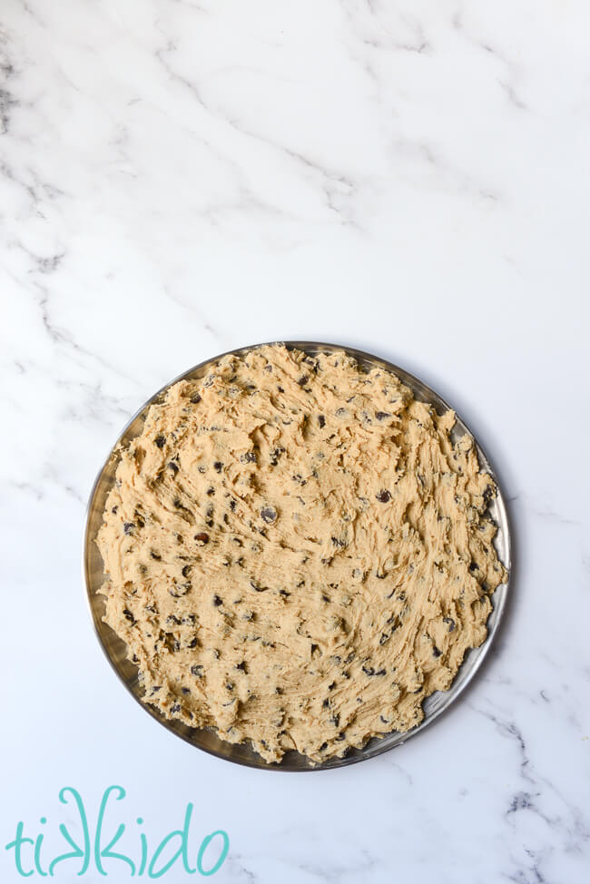 Chocolate chip cookie dough spread in a 12" pizza pan to make a cookie pizza, on a white marble surface.