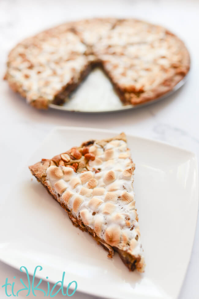 Slice of Rocky Road pizza cookie on a white plate, with the rest of the cookie pizza on a pizza pan in the background.