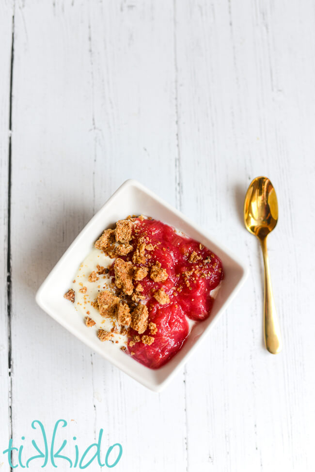 Square white bowl filled with greek yogurt, stewed rhubarb, and granola, next to a golden spoon on a white wooden surface.