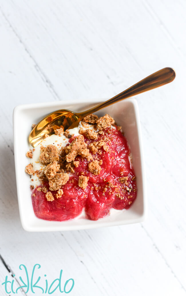 Small white bowl with stewed rhubarb, greek yogurt, and granola, with a golden spoon.