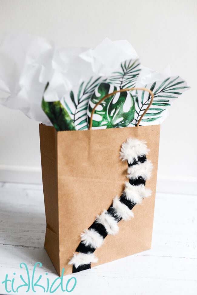 Lemur Gift Bag with a furry lemur tail decorating the front of the bag and paper tropical leaves and tissue paper peeking out of the top of the bag.