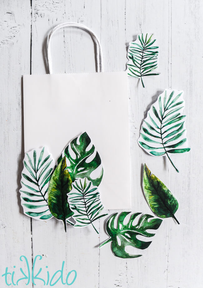 Printable tropical leaves being arranged on a white gift bag to make a tropical gift bag.