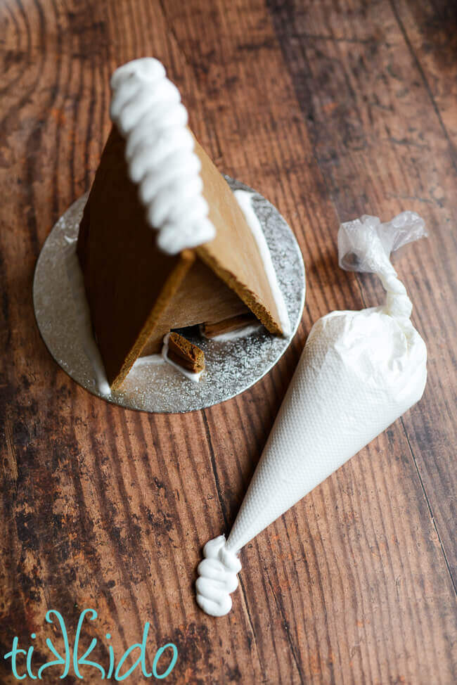 Eggless, vegan gingerbread house on a silver cake round, with a bag of vegan royal icing next to it on a dark wooden surface.