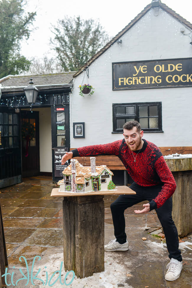 Delivering the gingerbread house Ye Olde Fighting Cocks to the actual pub in St Albans, England. A very happy man in a red jumper holds his hands in the air around the large gingerbread house.