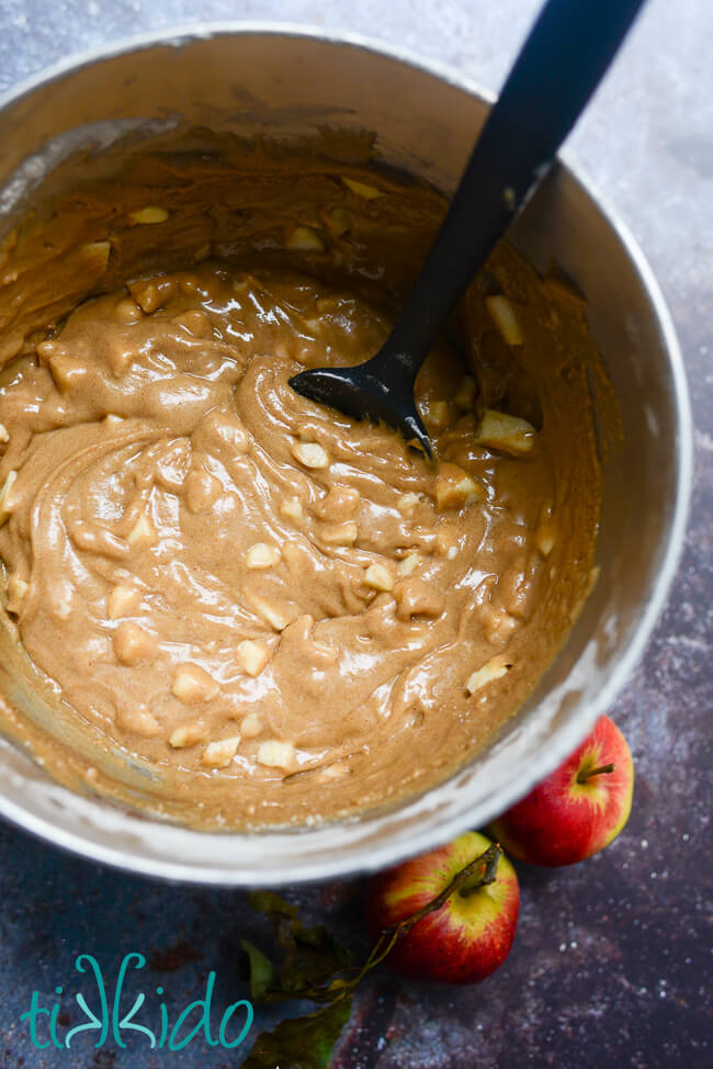 Thick apple quick bread batter strewn with chunks of fresh apple in a silver mixing bowl with a black spatula.