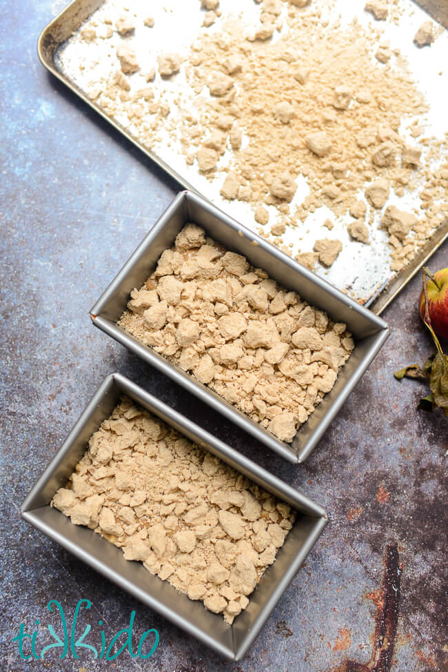 Apple quick bread batter divided between two bread pans and topped with crumb topping before baking.  A baking sheet with extra crumb topping sits at the top of the photo.