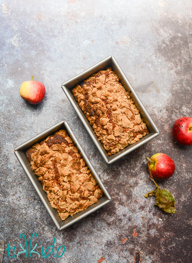 Two loaves of crumb topped apple bread, still in their pans, on a concrete surface, surrounded by three whole apples.