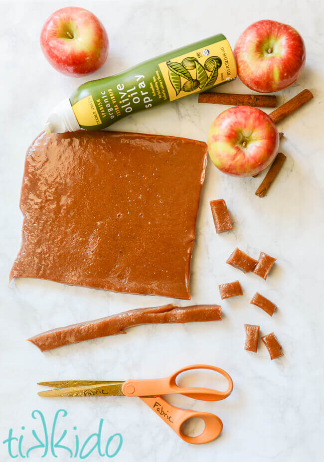 Apple cider caramels being cut into pieces, surrounded by scissors, apples, and olive oil spray.