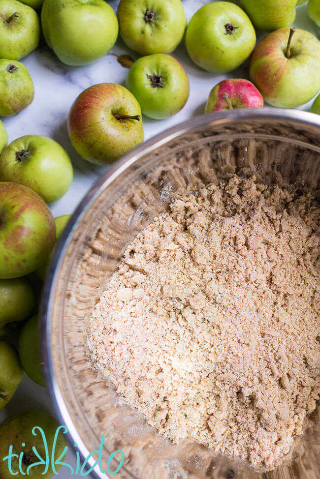 Apple crumble topping mixture in a silver bowl, surrounded by green apples.