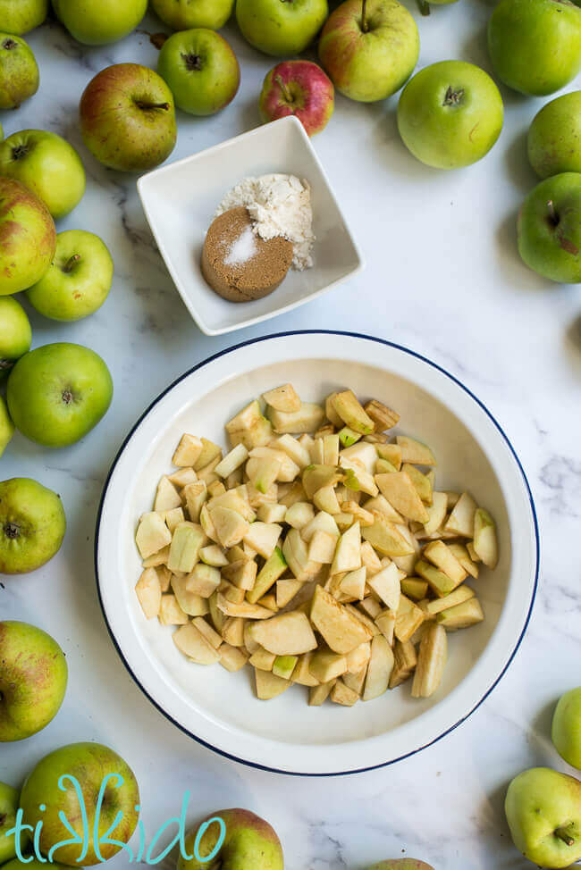 Sliced apples in a pie plate, next to ingredients for apple crumble filling, surrounded by green apples.