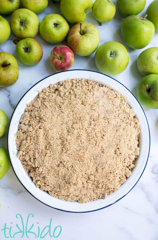 Unbaked apple crumble in a pie tin, surrounded by fresh apples.