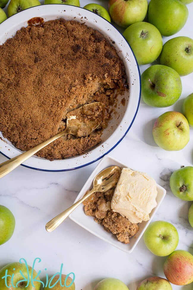 Apple crumble baked in a pie tin, and scooped into a white bowl, surrounded by green apples.