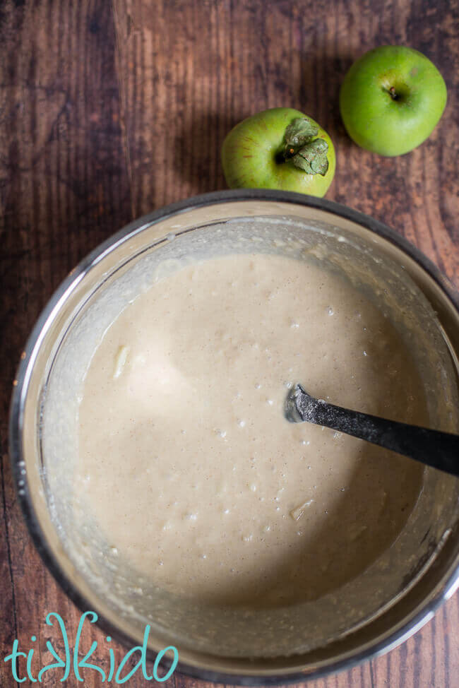 Batter for apple pancakes recipe in a silver bowl, sitting on a wooden table.