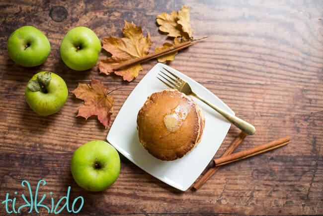 Apple pancakes on a white plate, sitting on a wooden table, surrounded by fresh apples, cinnamon sticks, and fall leaves.
