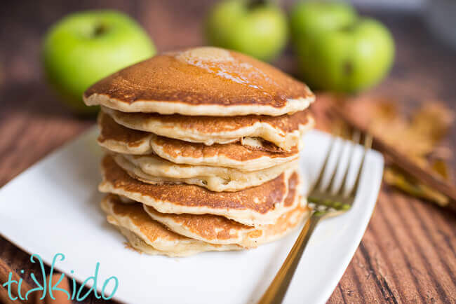 Stack of apple cinnamon pancakes with a pat of butter melting on top, and fresh apples in the background.