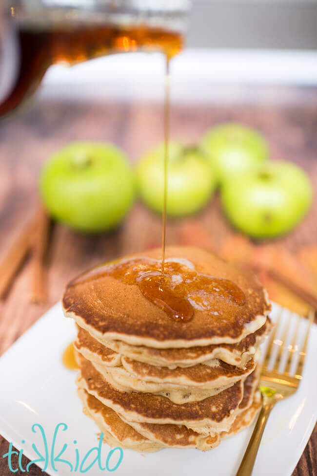 Maple syrup being poured on a stack of apple cinnamon pancakes.