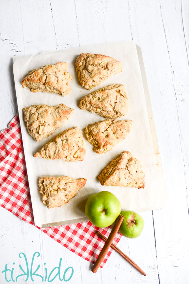 Freshly baked apple scones on a parchment lined baking sheet, sitting on a white wooden surface next to two green apples and two cinnamon sticks.