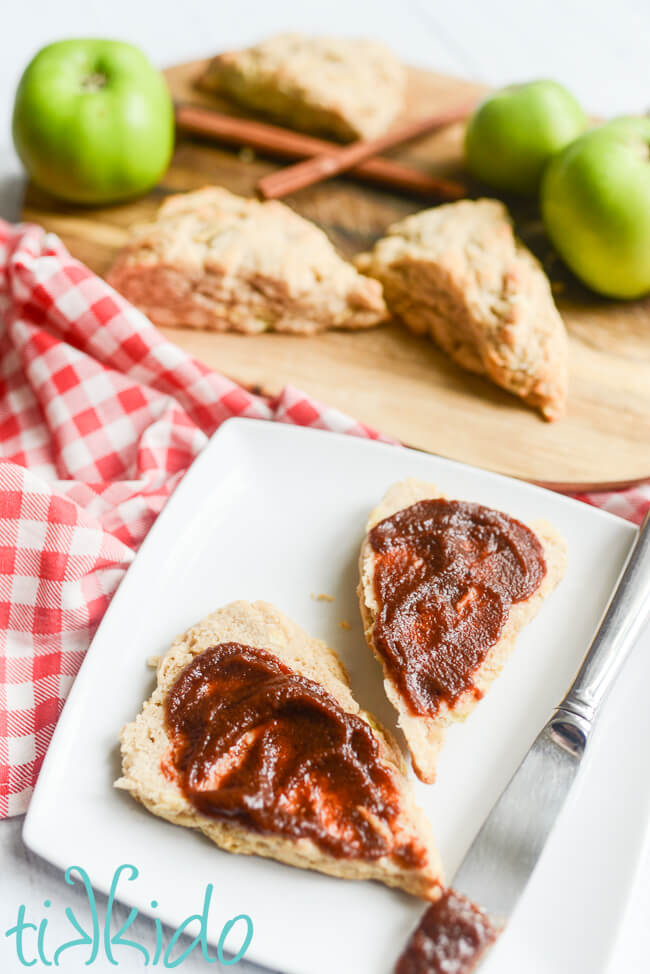 White plate with an apple scone split in half and spread with apple butter, with a cutting board with more apple scones and fresh green apples in the background.