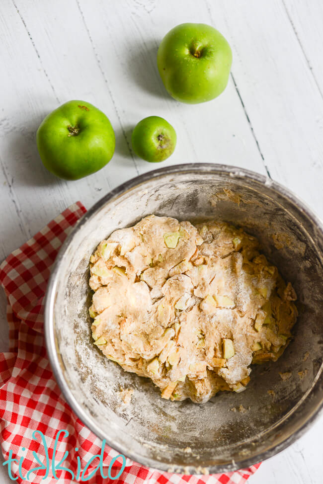 Silver bowl full of dough for Apple Scones, sitting on a white wooden surface with a red gingham kitchen towel and fresh green apples.