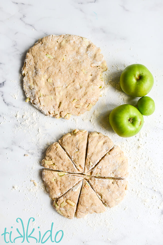 Apple scone dough divided into two 8" disks on a floured marble surface.  One of the disks is cut into eight equal wedges.  Three green apples sit next to the dough.