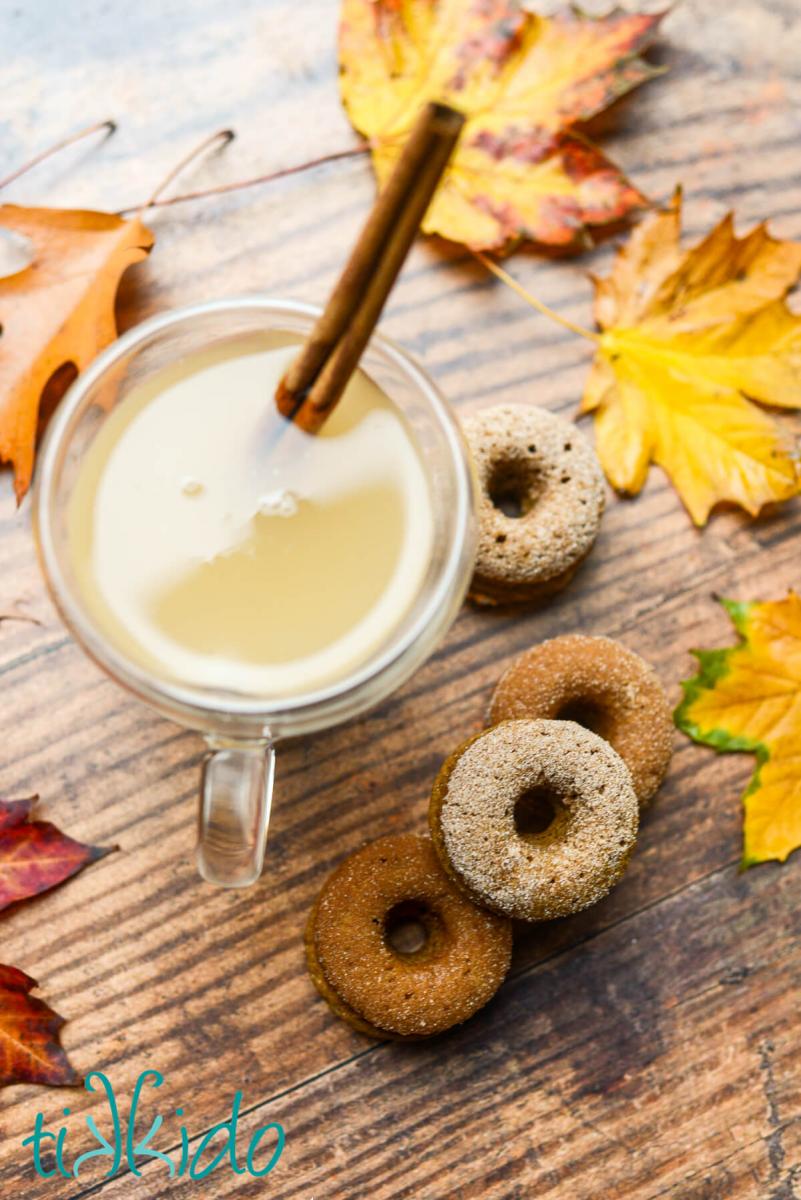 Overhead shot of four mini apple cider donuts on a wooden surface next to a glass mug full of apple cider.  Colorful fall leaves are scattered around on the dark brown table surface.