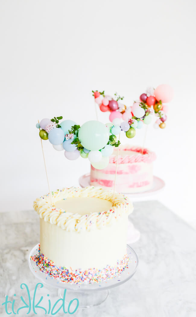 Two cakes decorated with American Buttercream Frosting on cake stands, topped with mini balloon garland cake toppers.