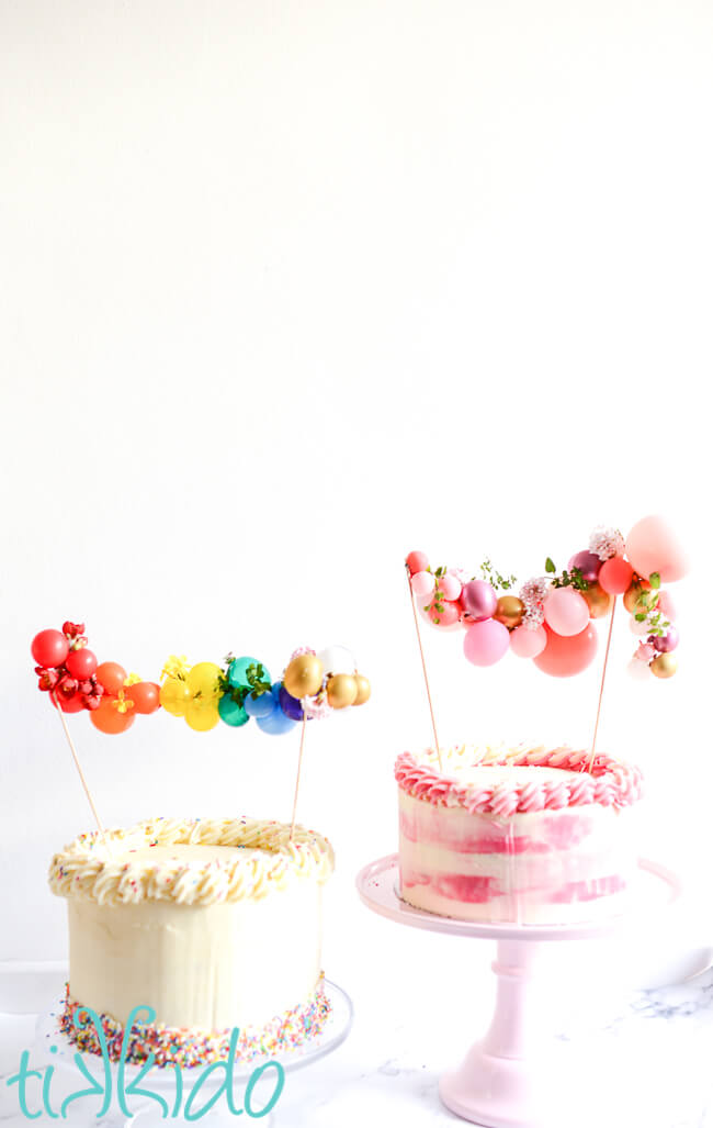 Two cakes, one with a rainbow balloon garland cake topper, one with a pink mini balloon garland cake topper.