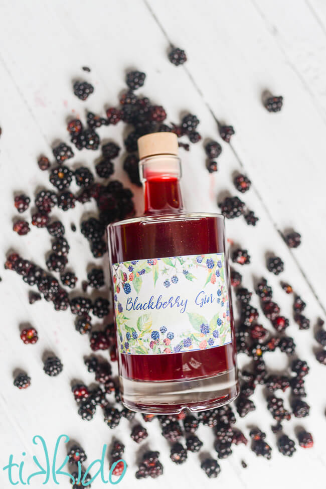 Bottle of blackberry gin surrounded by fresh blackberries, on a white wooden surface.