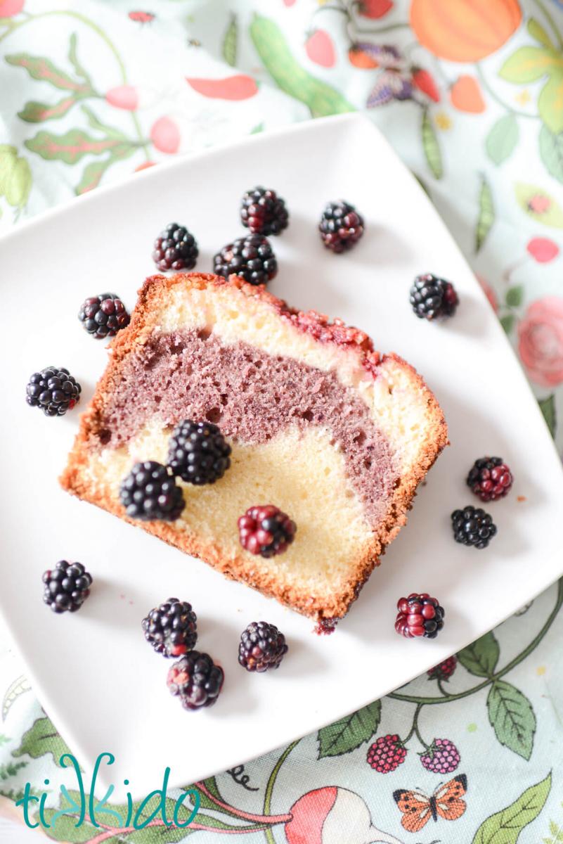 Blackberry Pound Cake Recipe Slice of blackberry pound cake on a square white plate, with fresh blackberries scattered around the slice of cake. The plate sits on a kitchen towel printed with designs of fresh berries and garden vegetables.