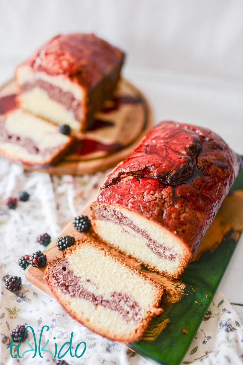 Blackberry Pound Cake Recipe Two loafs of blackberry pound cake on two cutting boards. Both have a slice of the pound cake cut to reveal the blackberry swirl inside of the cake. Fresh blackberries are scattered around the cakes.