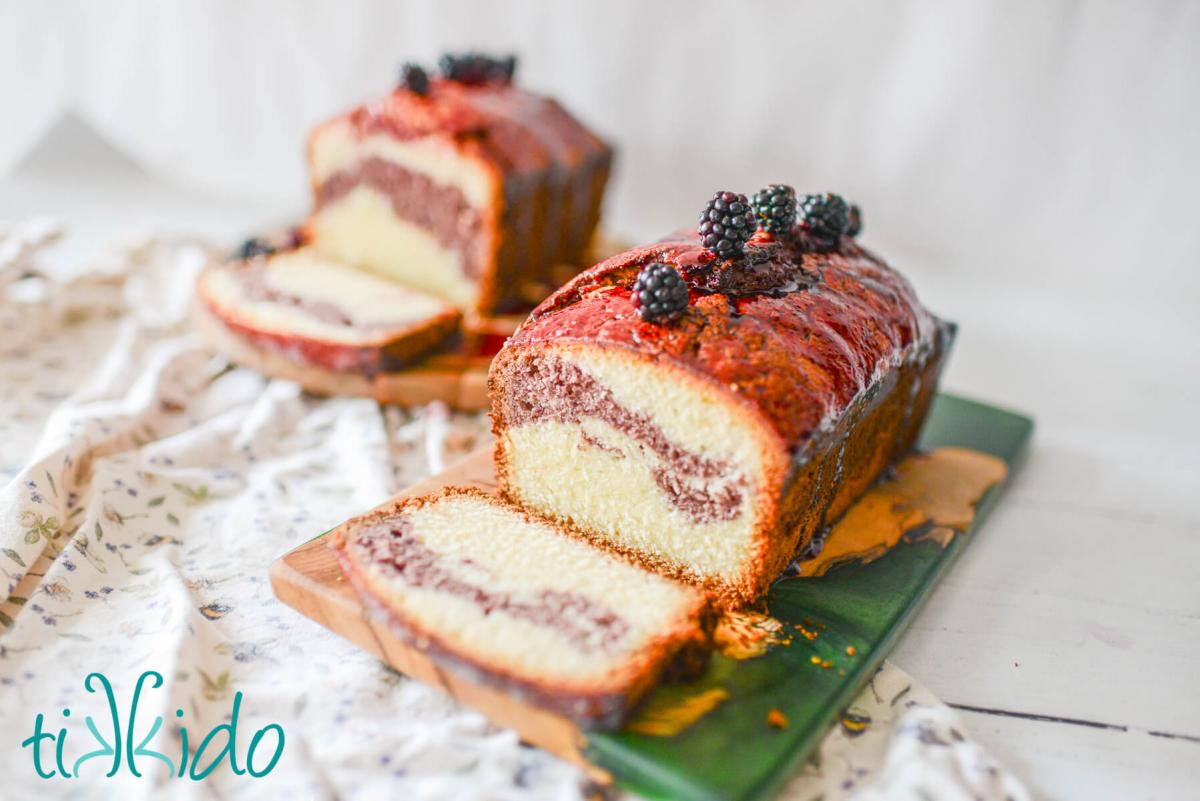 Blackberry Pound Cake Recipe Two loaves of blackberry pound cake sitting on two wooden cutting boards. A slice is cut out of each loaf to show the blackberry swirl inside the cake, and the tops of the cakes are decorated with a line of fresh blackberries.
