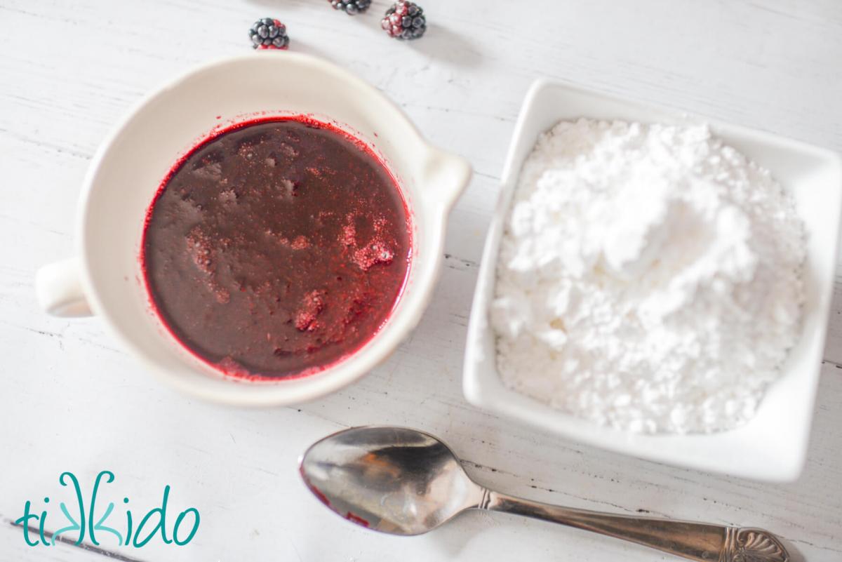 Blackberry Pound Cake Recipe Cup with fresh blackberry puree and a small bowl of powdered sugar sitting on a white wooden surface next to a spoon, ready to make blackberry glaze.