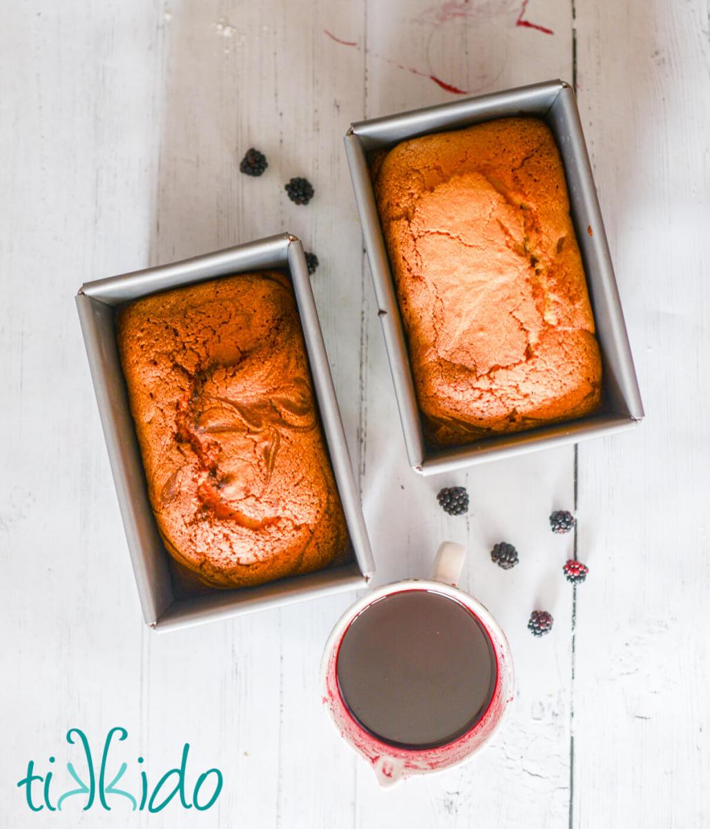 Blackberry Pound Cake Recipe Two loaves of freshly baked blackberry pound cake and a cup of blackberry glaze on a white wooden surface.