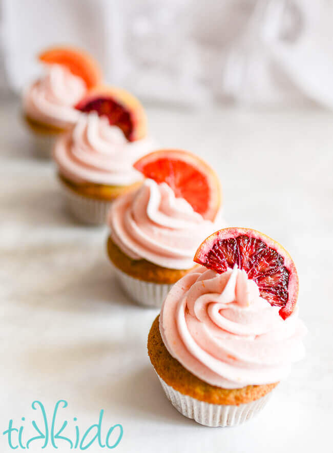 Four blood orange cupcakes topped with naturally pink blood orange frosting and slices of blood orange on a white background.