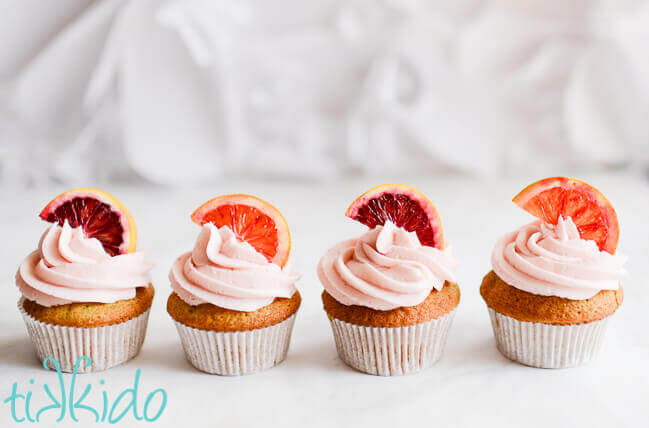 Four blood orange cupcakes topped with naturally pink blood orange frosting and slices of blood orange on a white background.