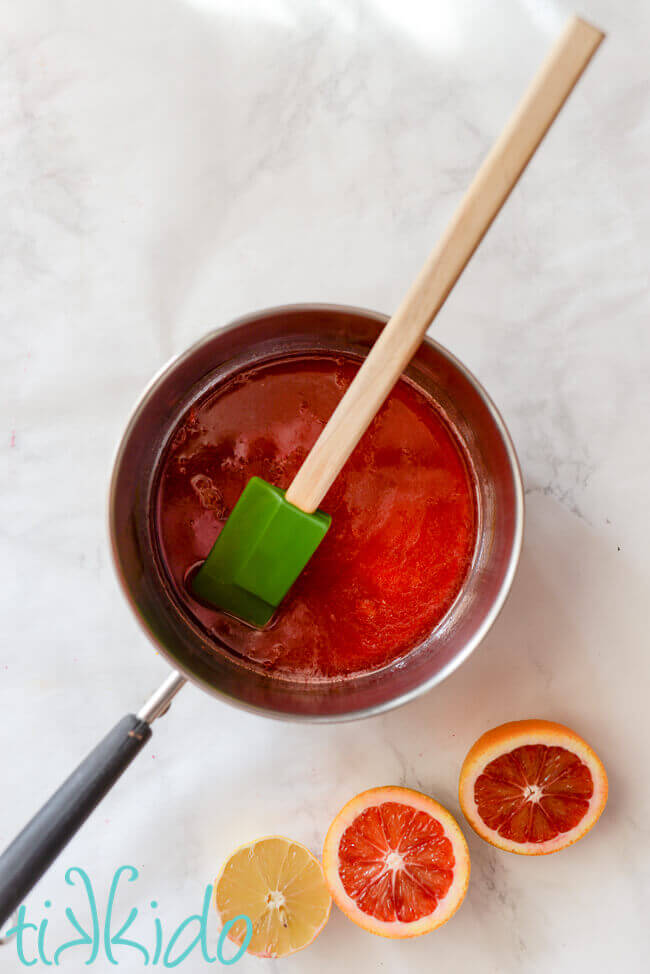 Blood orange simple syrup in a small saucepan on a white marble surface, next to sliced blood oranges and lemon.