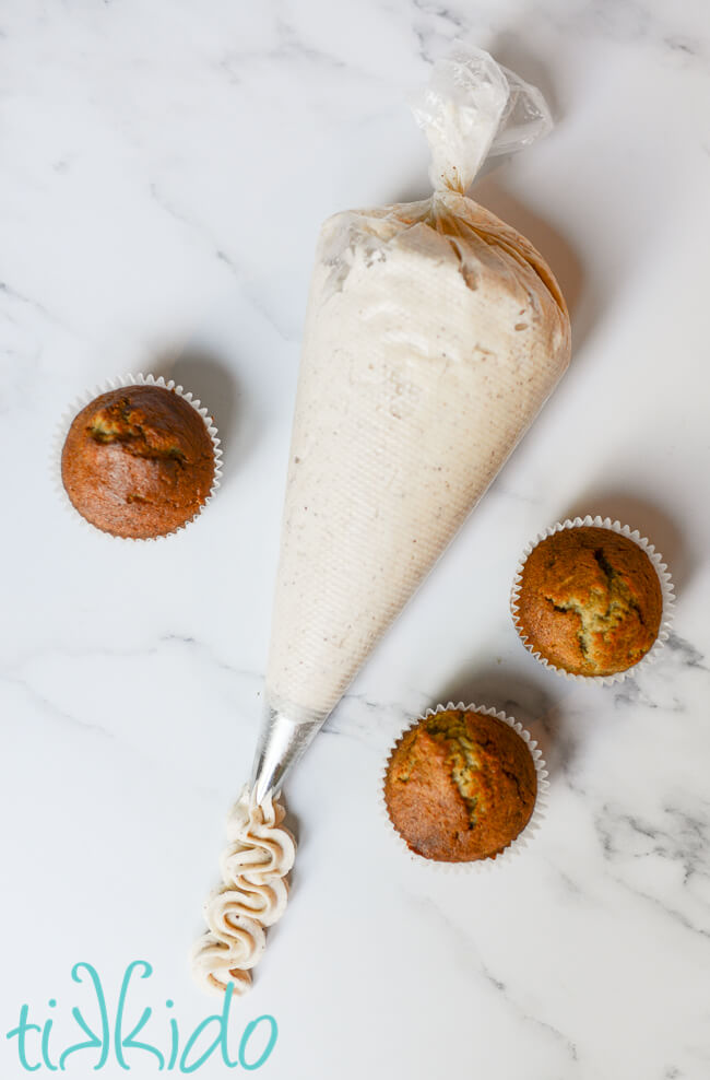 Icing bag of Browned Butter Frosting on a white marble surface, surrounded by three banana cupcakes.