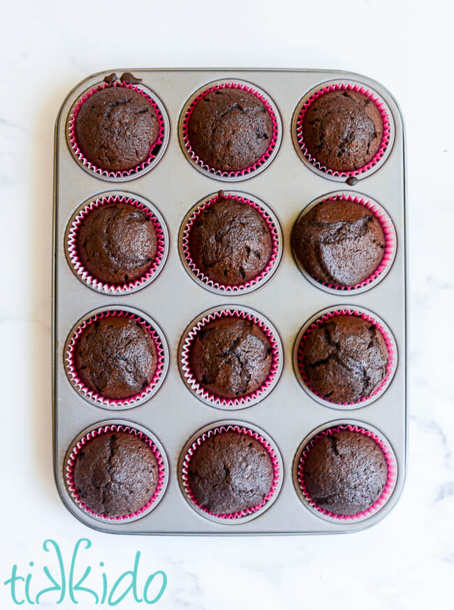 Cupcake tin filled with 12 freshly baked chocolate cupcakes on a white marble surface.